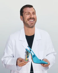 Smiling man wearing white coat holding a bright blue slingback shoe against a pale background