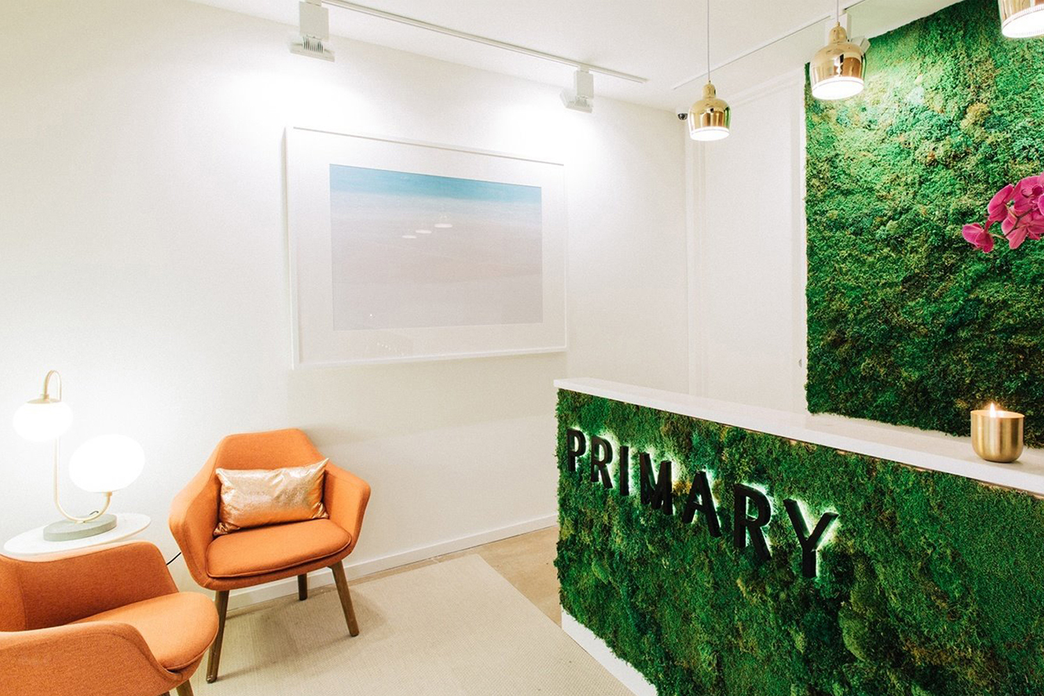 Modern office reception with a moss wall, backlit primary lettering desk, and orange armchairs designed by DIA Studio.