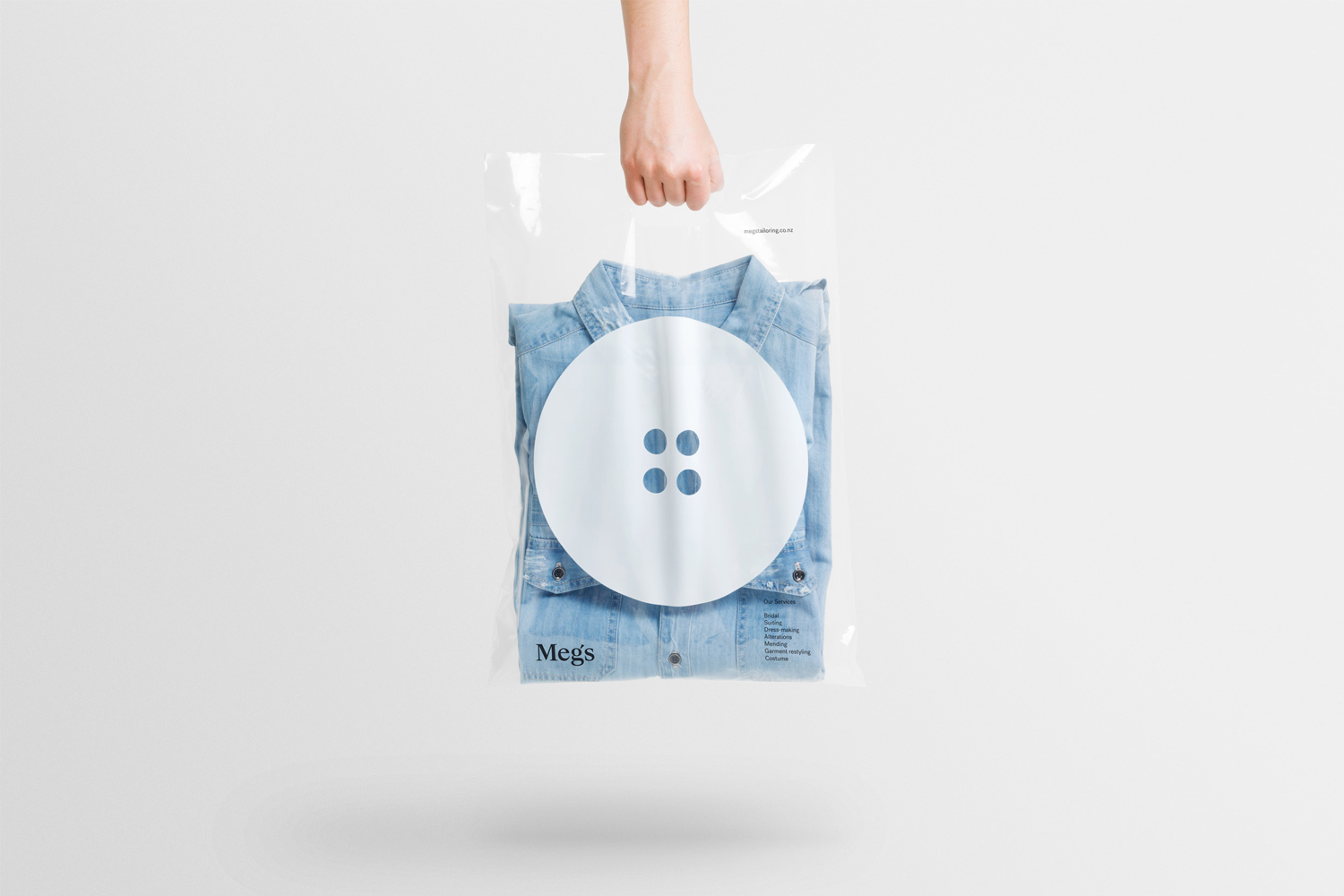 Hand holding a clear plastic garment bag containing a light blue denim shirt against a white seamless background
