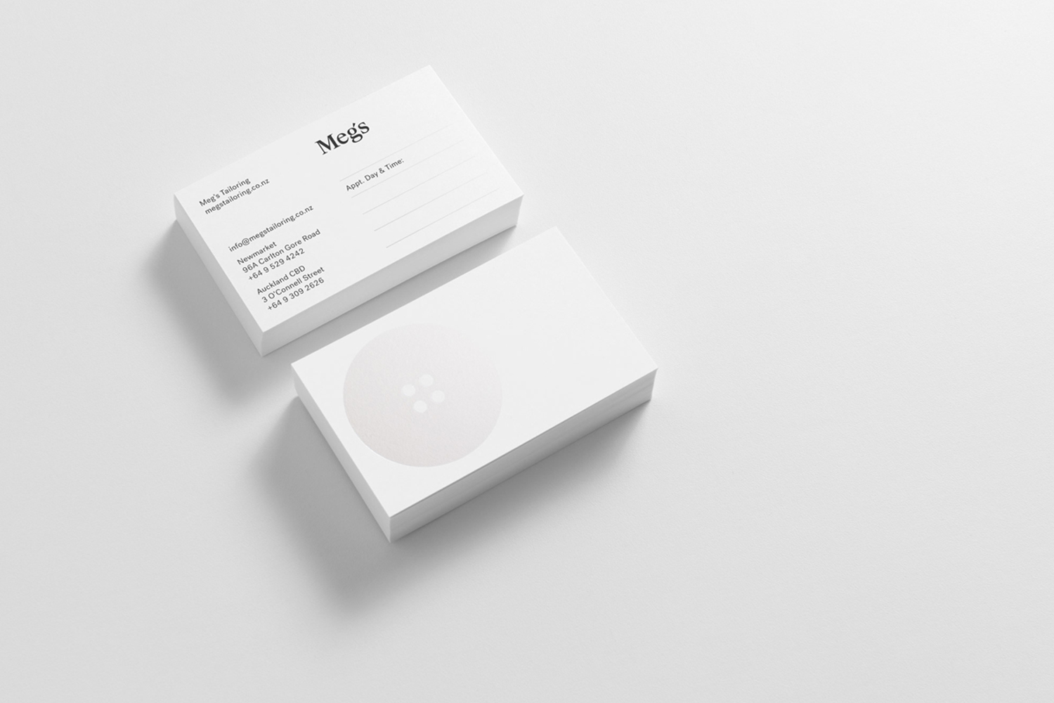 Two white business cards for Meg's Tailoring stacked on a clean light grey surface, showing typography and a subtle button graphic