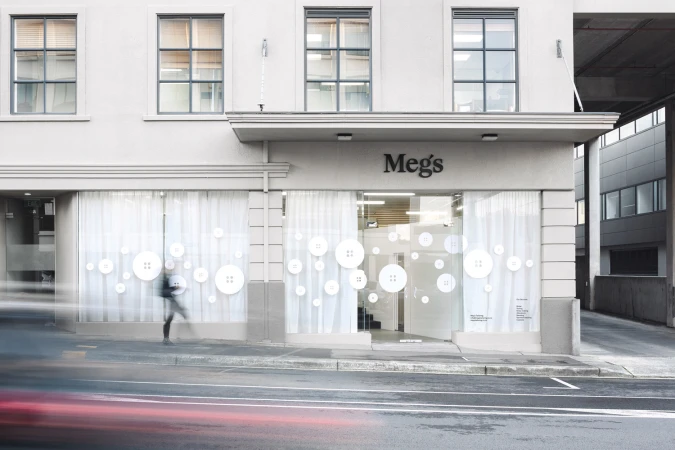 Storefront of Meg's Tailoring featuring large white button decals on windows, exterior signage, and passing traffic.