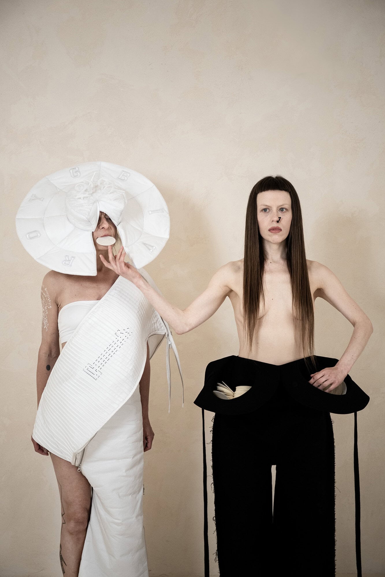 Two models showcasing avant-garde white and black sculptural apparel against a plain textured wall