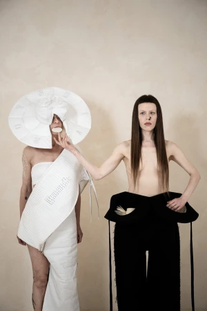 Two models showcasing avant-garde white and black sculptural apparel against a plain textured wall