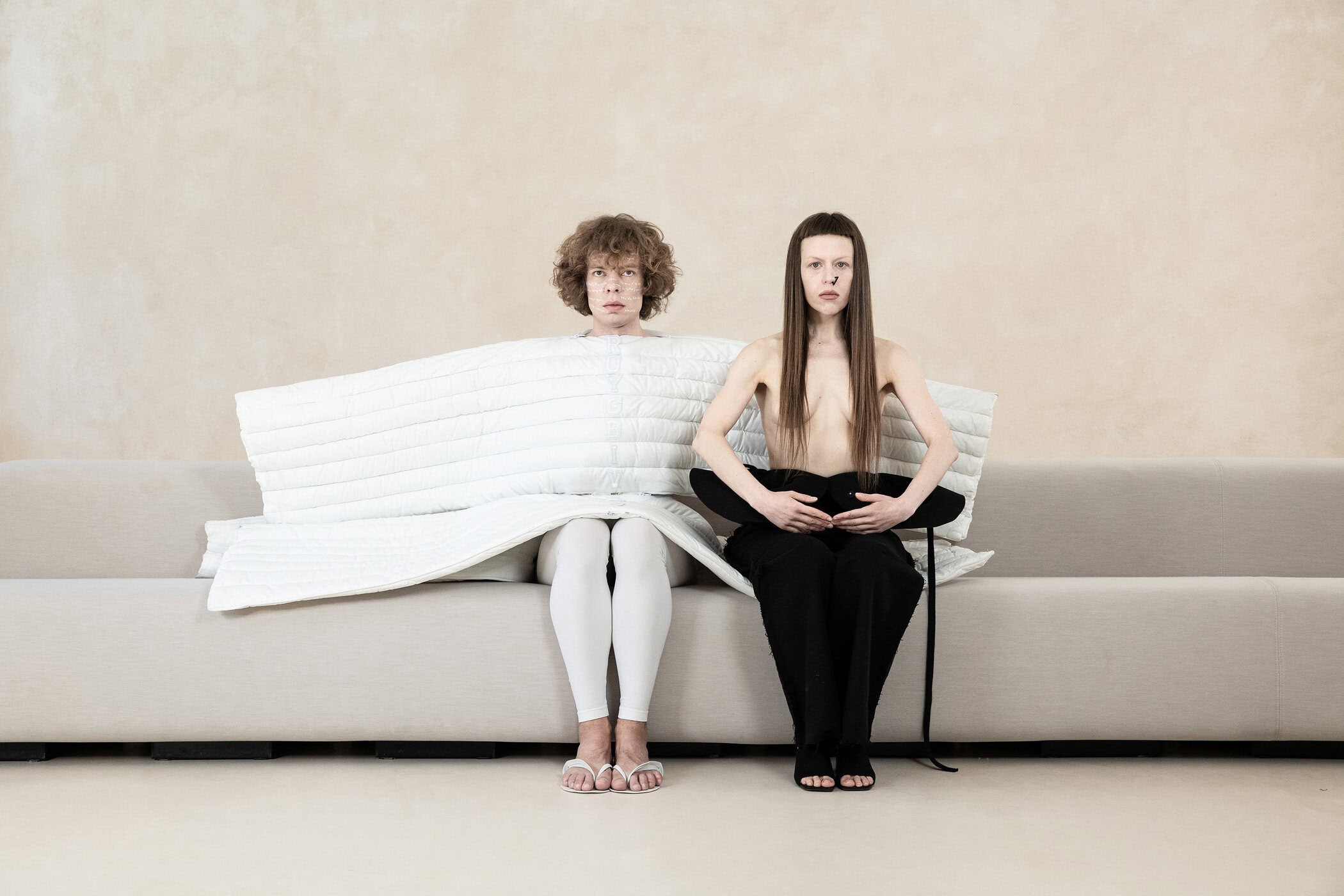 Two models seated on a long light grey sofa against a neutral textured wall for the Dzhus Anticon campaign.