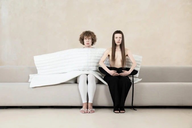 Two models seated on a long light grey sofa against a neutral textured wall for the Dzhus Anticon campaign.