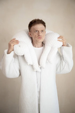 Man wearing a textured white coat with a large padded collar and writing on his forehead against a neutral background