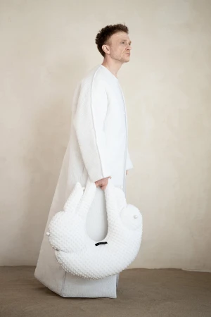 Man wearing a long white textured coat holding a sculptural white handbag against a neutral background