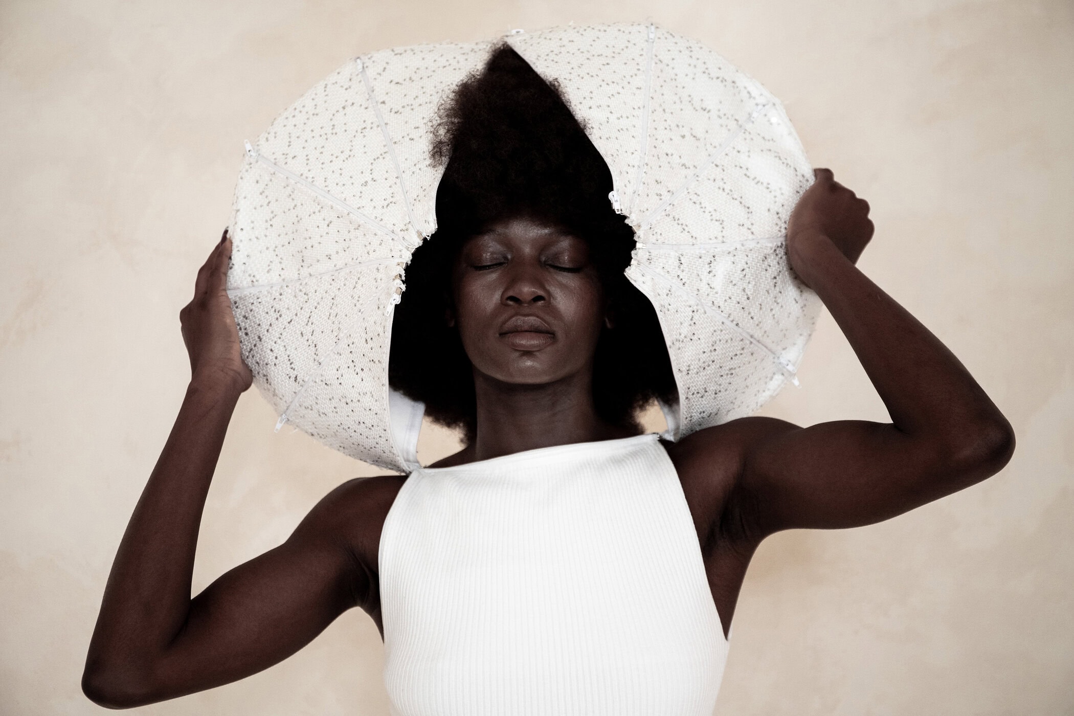 Model with eyes closed wearing a white sleeveless top holding a sculptural white circular garment around her head