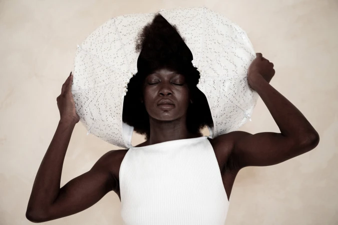 Model with eyes closed wearing a white sleeveless top holding a sculptural white circular garment around her head