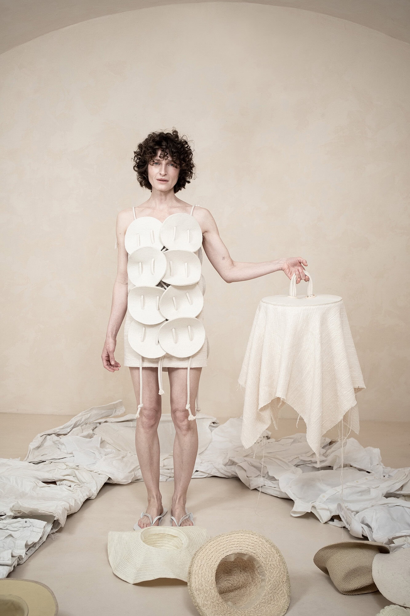 Model wearing a structured white sculptural garment holding a matching covered object, surrounded by hats on a neutral studio floor