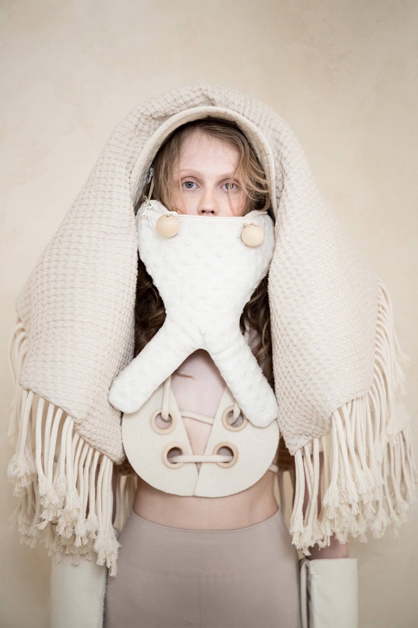 Model wearing sculptural cream apparel with draped fringe headpiece against a plain neutral backdrop for Dzhus Ad Campaign