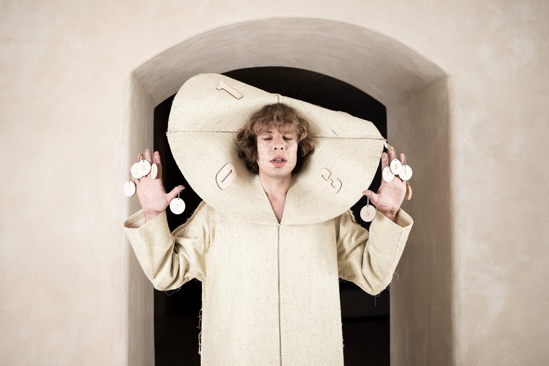 Model wearing a textured cream-colored sculptural clock-like hat and matching coat with numbered tokens dangling from fingers