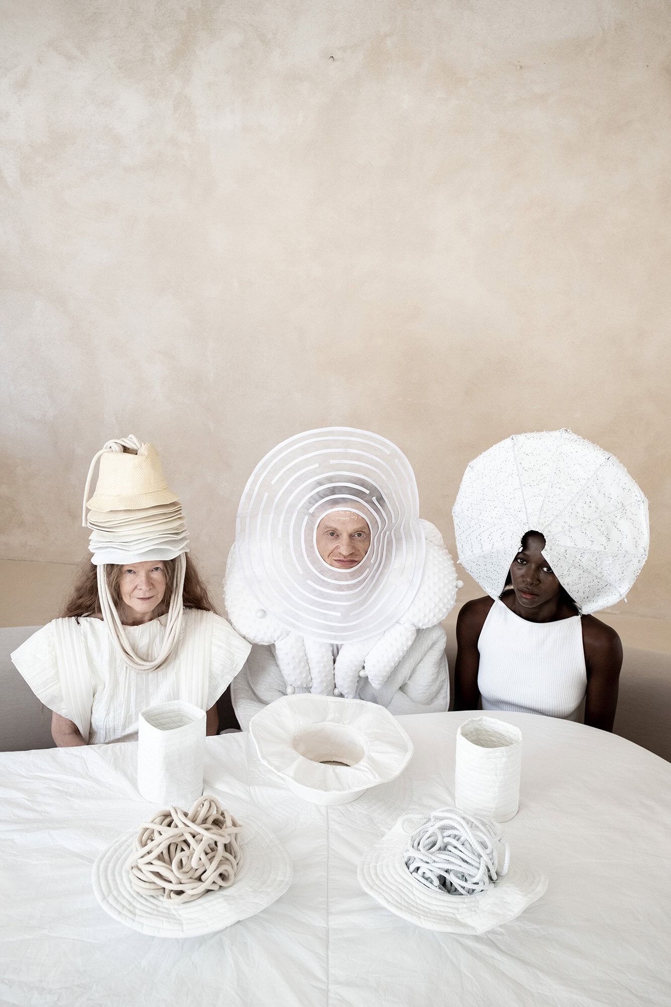 Three models wearing avant-garde sculptural white headwear and apparel seated around a white table against a textured neutral wall.