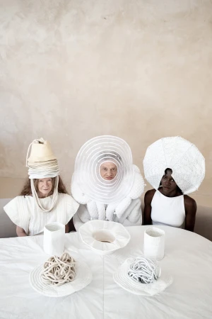 Three models wearing avant-garde sculptural white headwear and apparel seated around a white table against a textured neutral wall.
