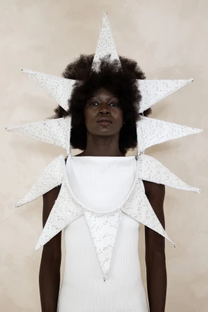 Black woman wearing a structured white star-shaped avant-garde headdress and matching garment against a textured background.