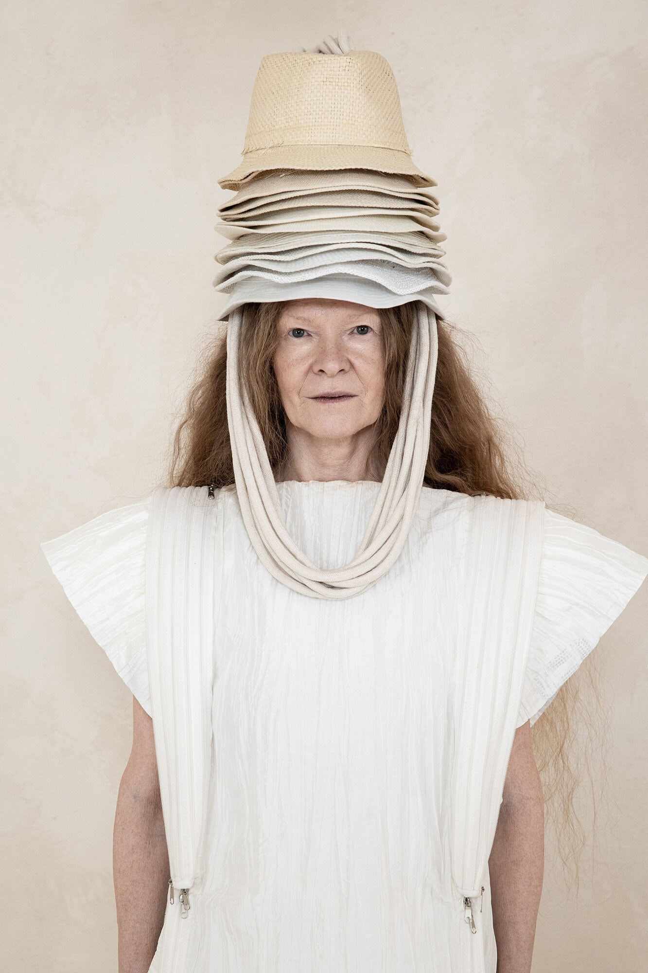 Model wearing a structured white dress and a tall stack of neutral hats on head for Dzhus Anticon campaign