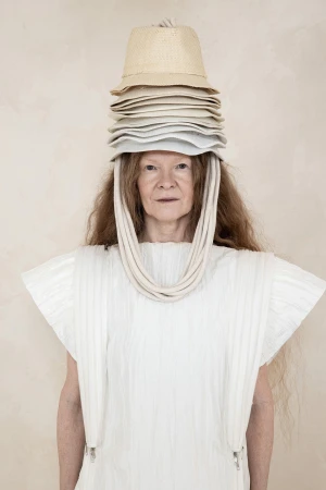 Model wearing a structured white dress and a tall stack of neutral hats on head for Dzhus Anticon campaign