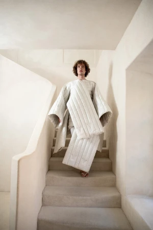 Model descending indoor white stairs wearing a structural quilted white coat for the Dzhus Anticon campaign