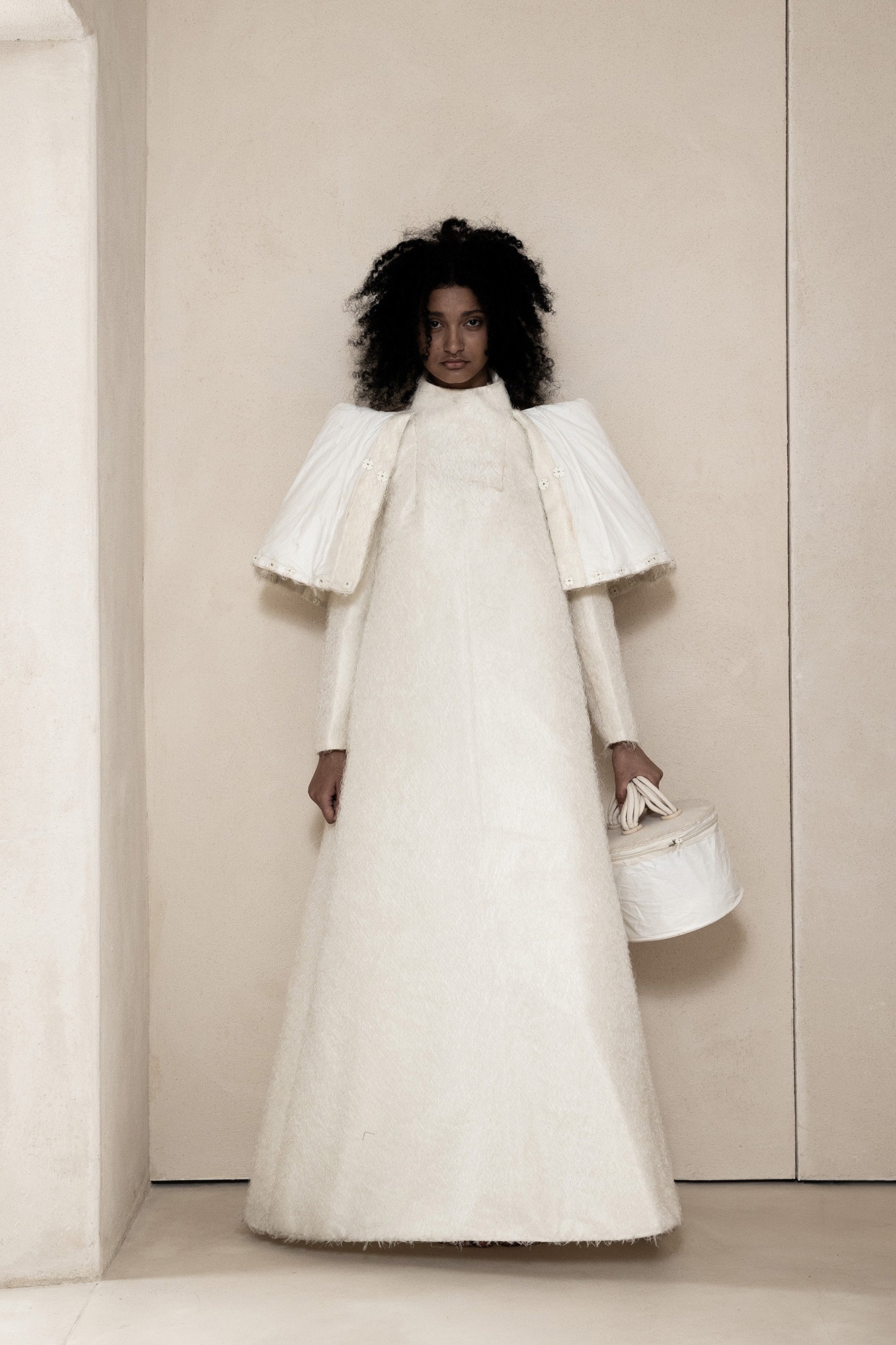 Model standing against a neutral wall wearing a structured white avant-garde gown with exaggerated shoulders and holding a cylindrical handbag.