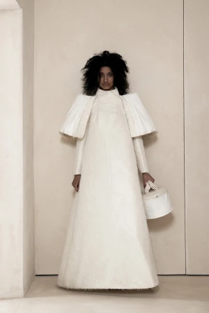 Model standing against a neutral wall wearing a structured white avant-garde gown with exaggerated shoulders and holding a cylindrical handbag.