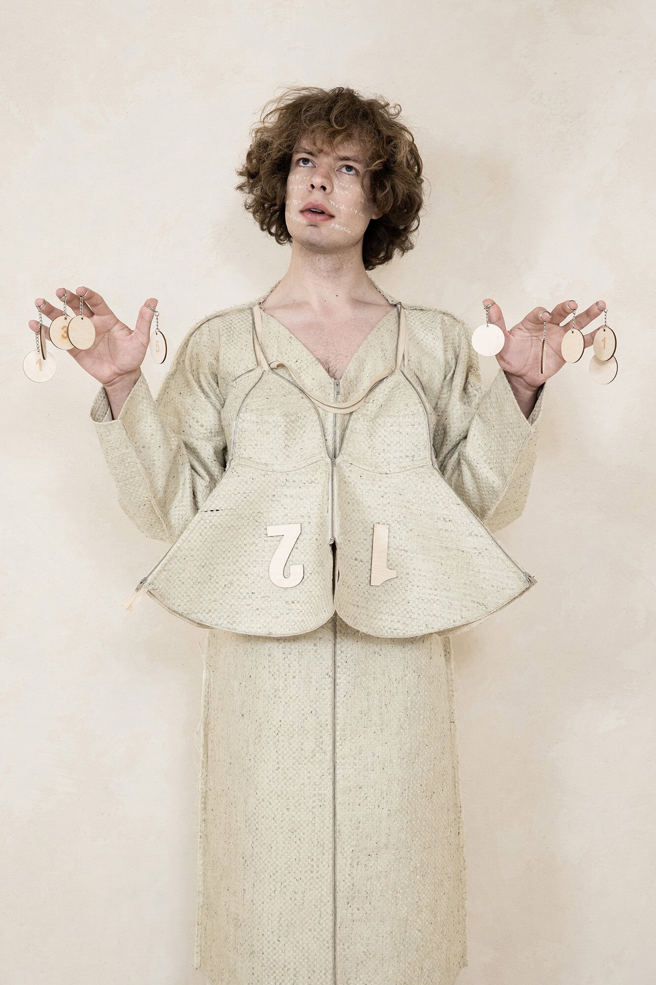 A man wearing an avant-garde structured beige garment holds small wooden discs against a textured neutral background