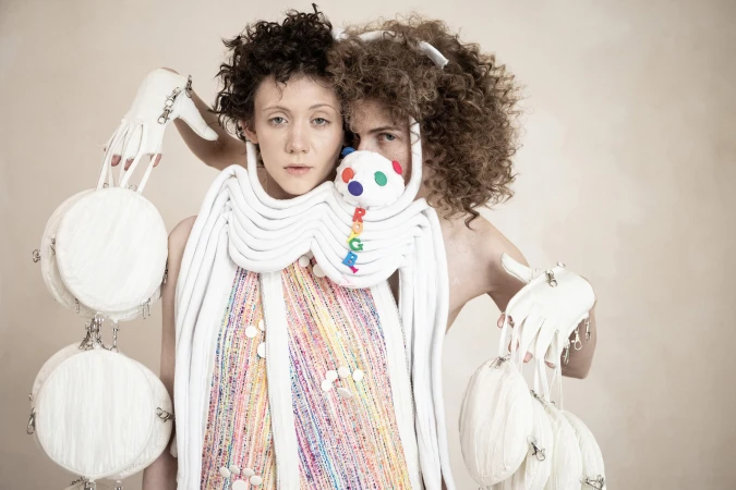 Two models wearing avant-garde white sculpted accessories and textured multi-colored garments against a plain neutral background.