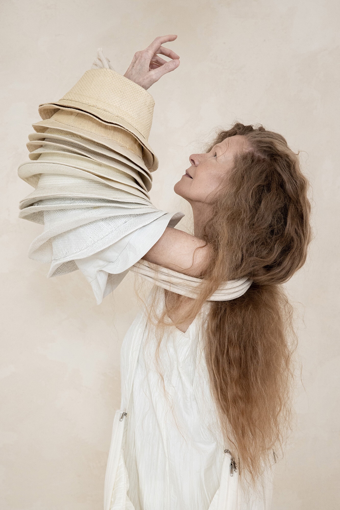 Woman wearing a stack of straw hats on her arm against a textured beige background