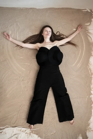 Woman in black sculptural outfit lying on sand with arms out, for Dzhus campaign
