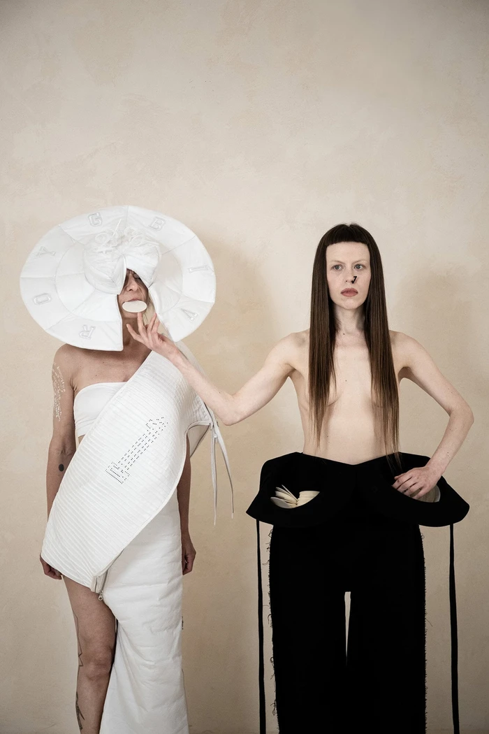 Two models showcasing avant-garde white and black sculptural apparel against a plain textured wall