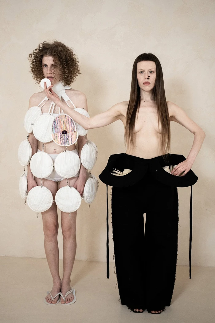 Two women modeling avant-garde avant-garde garments by Dzhus against a textured neutral background for the Anticon campaign.