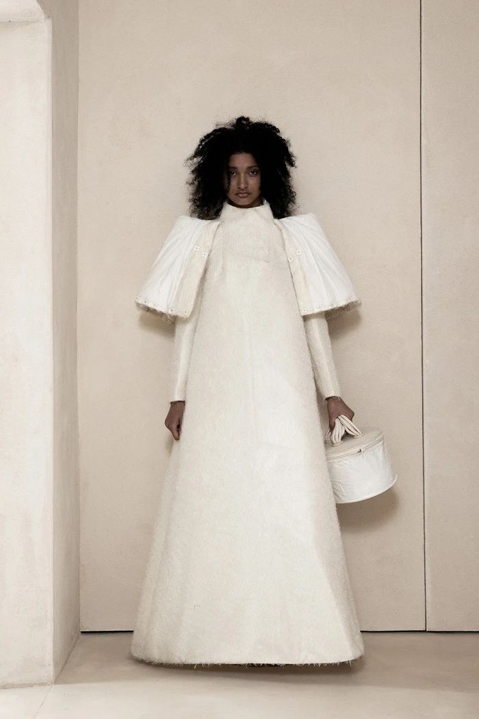 Model standing against a neutral wall wearing a structured white avant-garde gown with exaggerated shoulders and holding a cylindrical handbag.