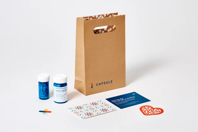 Kraft paper bag with Capsule branding, two small blue pill bottles, printed cards, and stickers on white seamless
