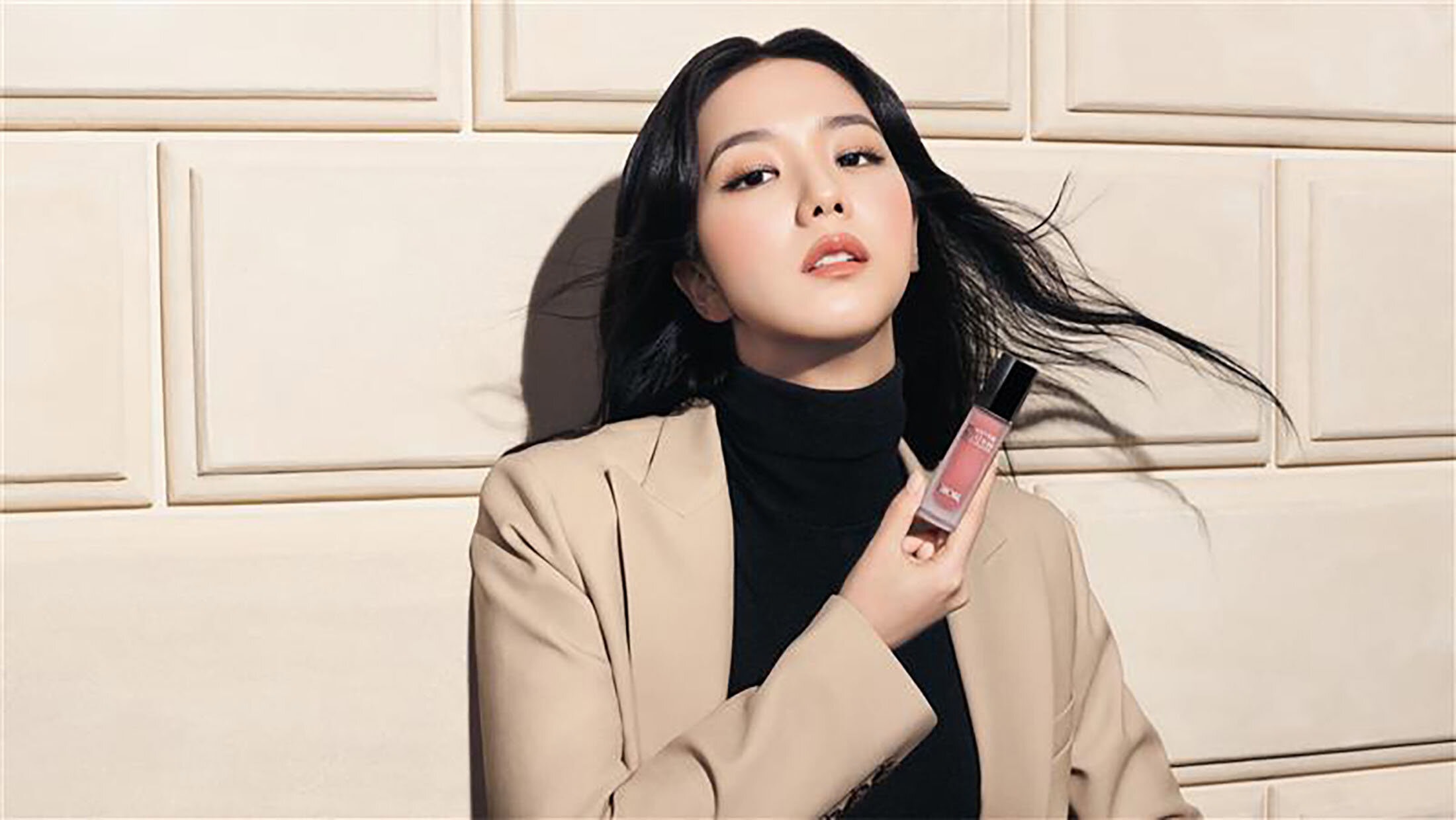 Jisoo holding a Dior makeup bottle while wearing a beige blazer and black turtleneck against a paneled wall