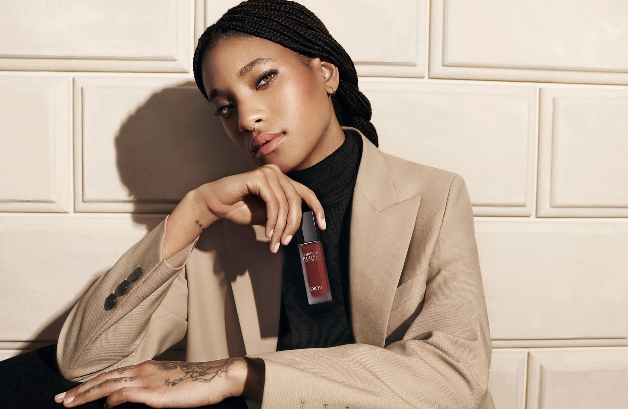 Willow Smith holding a Dior makeup product wearing a camel blazer against a panelled wall