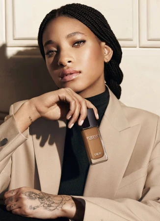 Willow Smith holding a Dior Forever foundation bottle wearing a beige blazer against a panelled wall