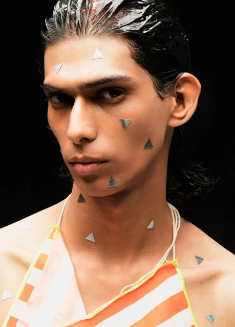 Portrait of a young adult male with wet dark hair and silver triangle appliques on his face, wearing a striped top against black.