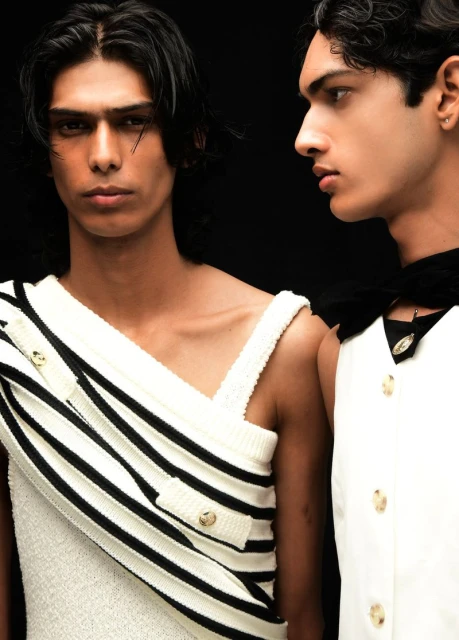 Two male fashion models pose in a studio against a dark backdrop, one wearing a white dress with black stripes and gold buttons.
