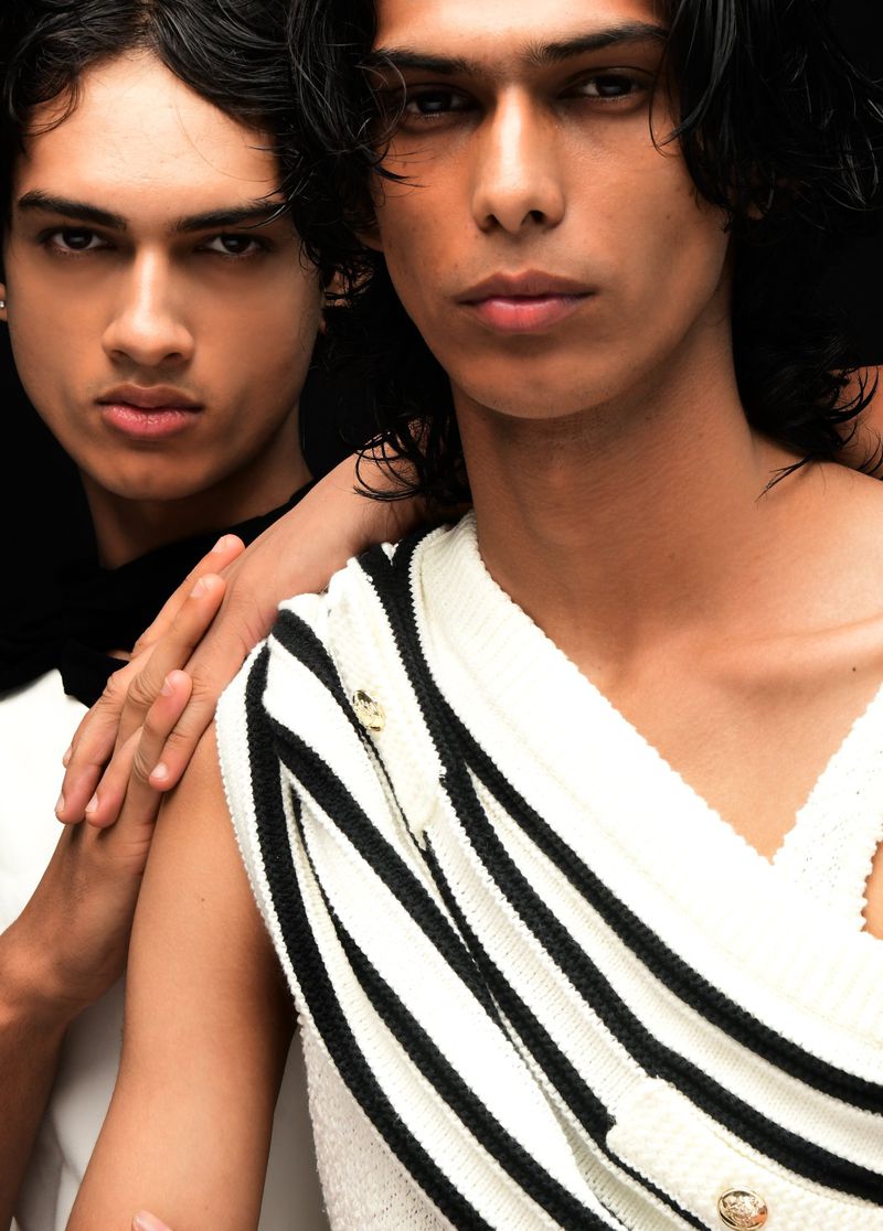 Close-up of two young adult male fashion models wearing striped knitwear in a studio setting with a dark background.
