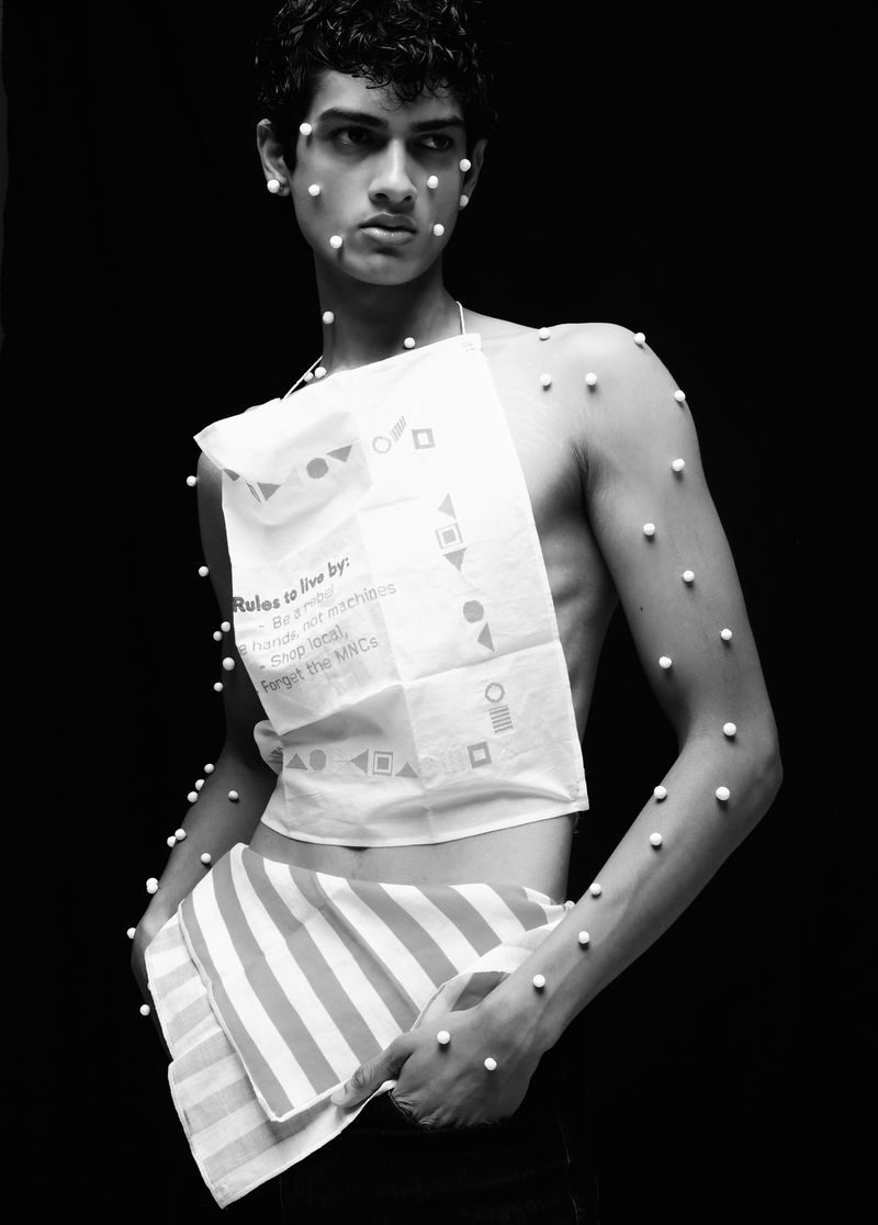 Artistic black and white portrait of a young man with beads on his face and body, wearing a patterned white cloth.