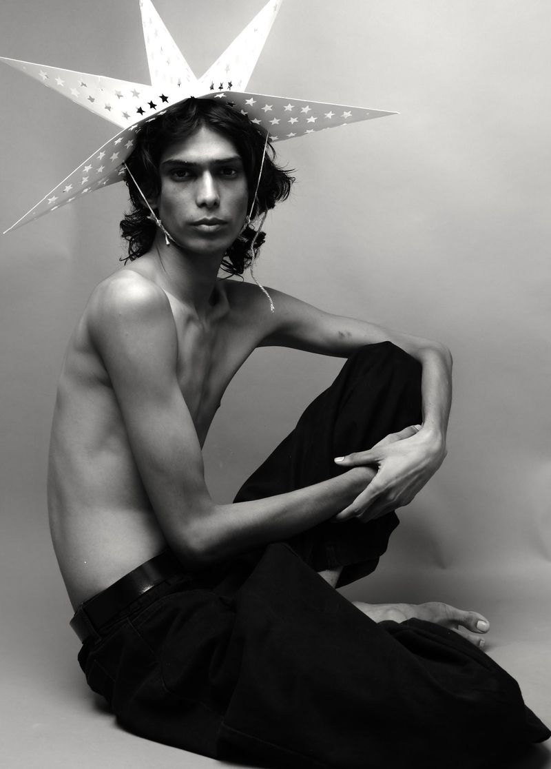 Monochrome portrait of a shirtless young adult male with long hair, wearing a star-shaped hat, and seated against a plain studio backdrop.
