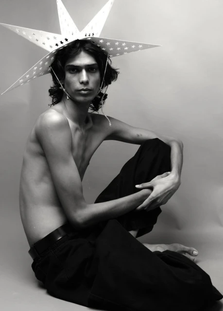 Monochrome portrait of a shirtless young adult male with long hair, wearing a star-shaped hat, and seated against a plain studio backdrop.