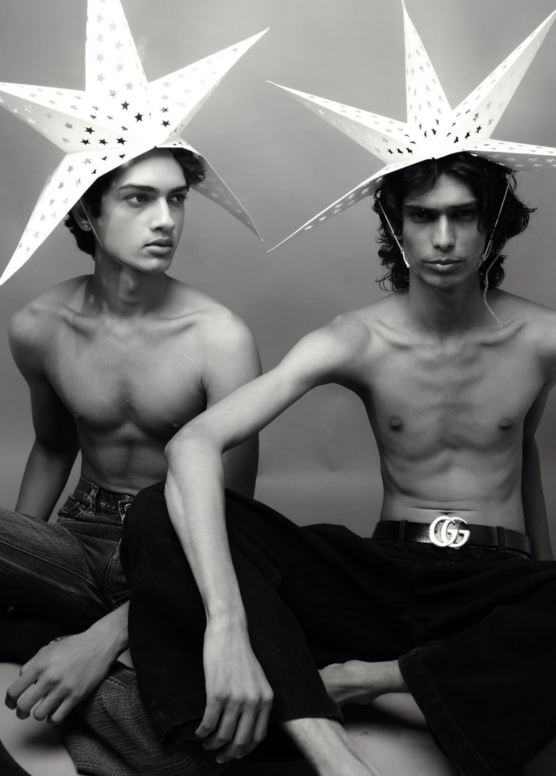 Two shirtless young men pose in a monochrome photo, wearing star-shaped hats, with one displaying a Gucci belt, against a neutral backdrop.