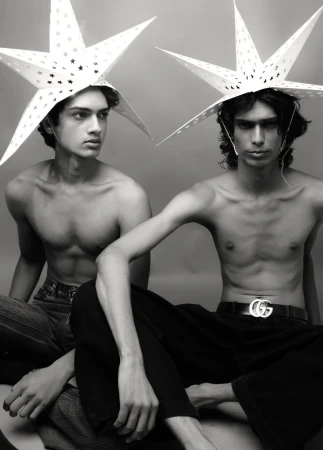 Two shirtless young men pose in a monochrome photo, wearing star-shaped hats, with one displaying a Gucci belt, against a neutral backdrop.