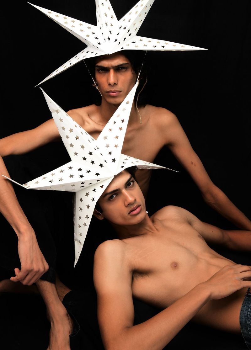 Two shirtless male models pose in a studio against a black backdrop, each wearing a large white star hat, creating a surreal, artistic image.
