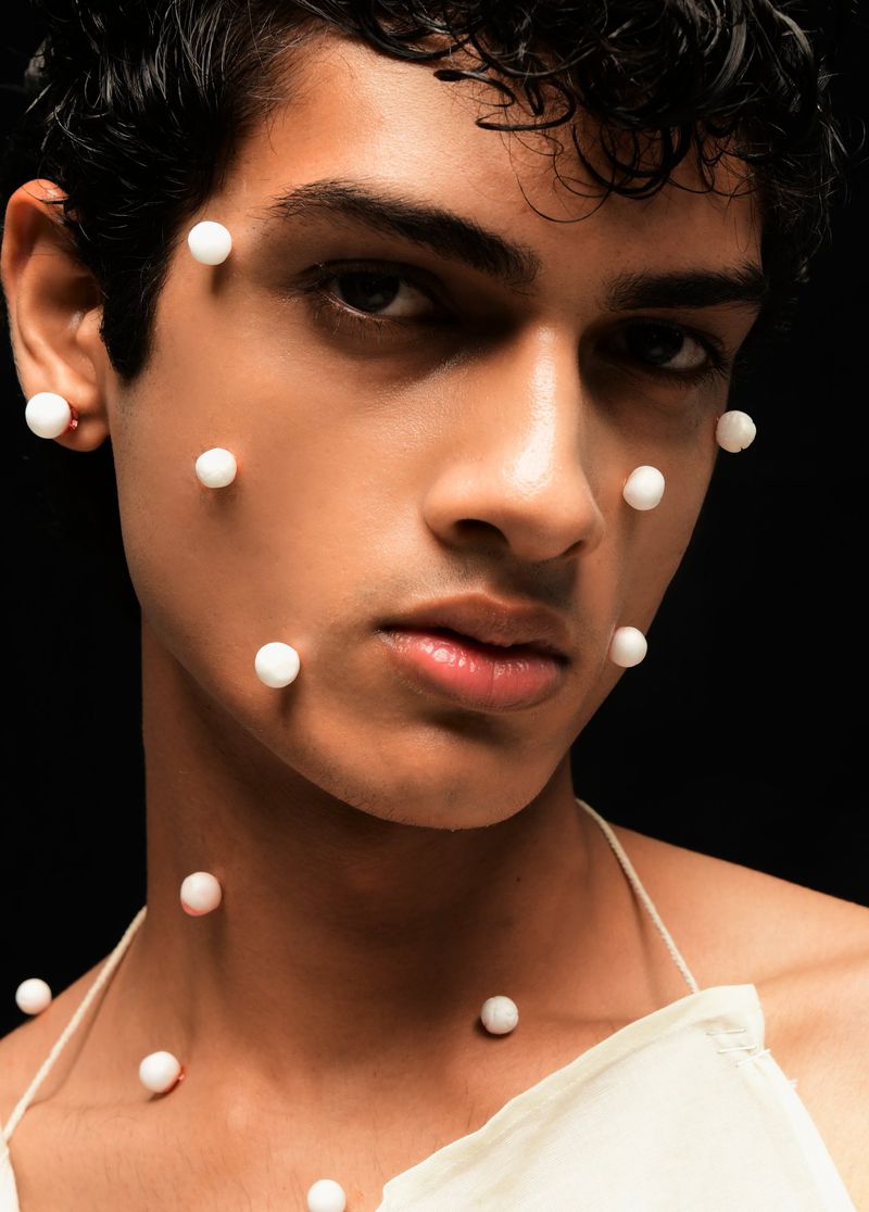 Close-up studio shot of a young adult male model with white spherical decorations adhering to his face and upper chest, against a black backdrop.