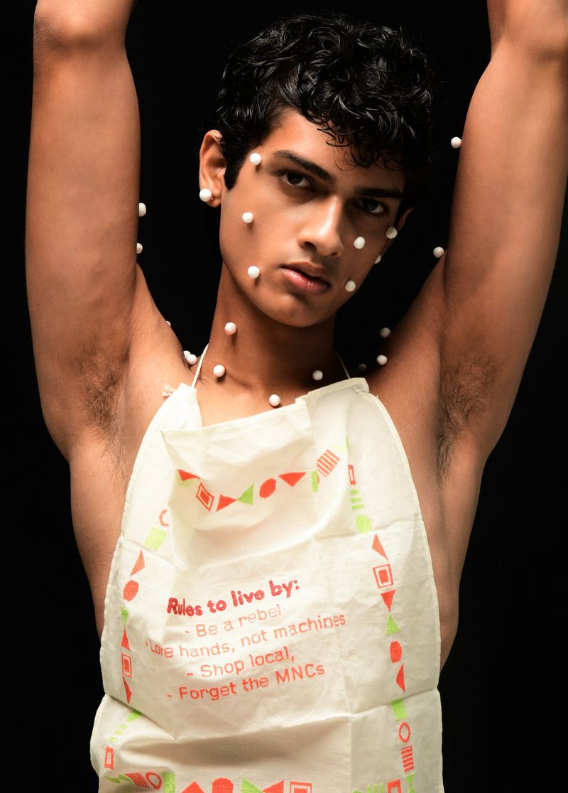 Young man poses with small white spheres on his face and arms, wearing a printed apron against a black background in a studio setting.