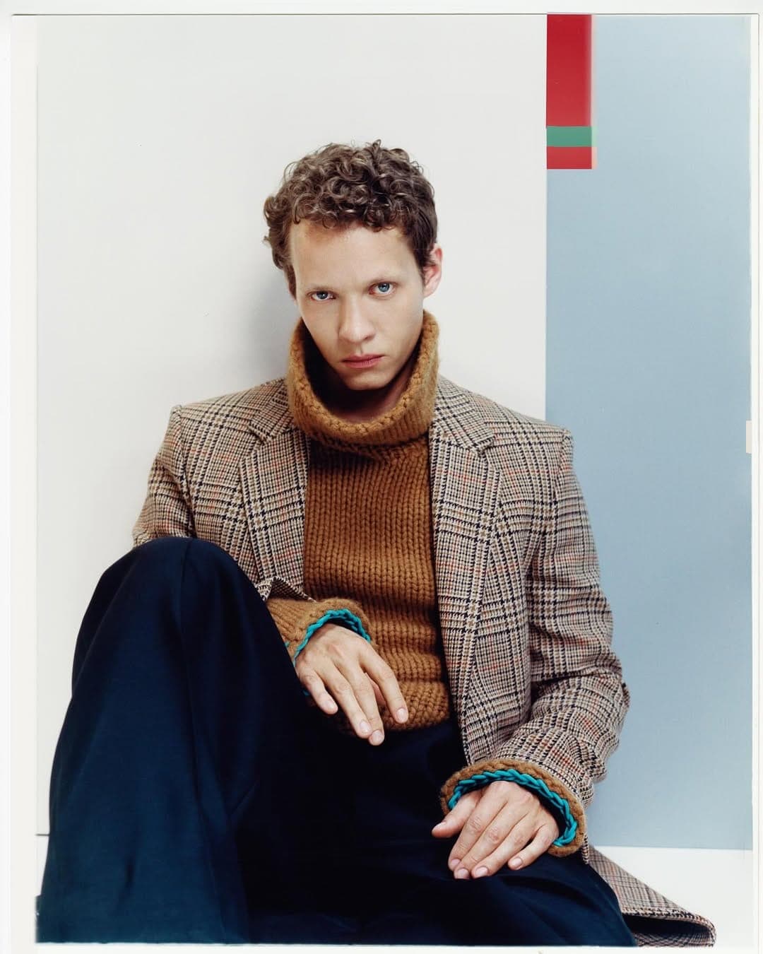 Man wearing a brown plaid blazer and turtleneck sweater seated against a geometric white and blue studio background