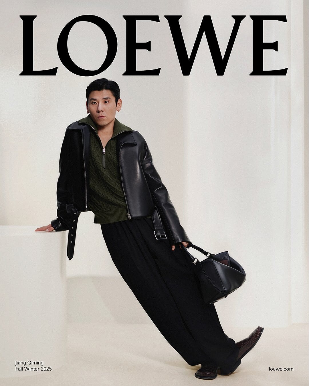 Man wearing a black leather jacket and olive green sweater leaning against a white pedestal, holding a black handbag.