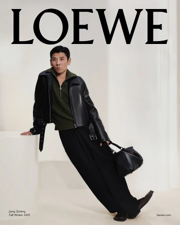 Man wearing a black leather jacket and olive green sweater leaning against a white pedestal, holding a black handbag.