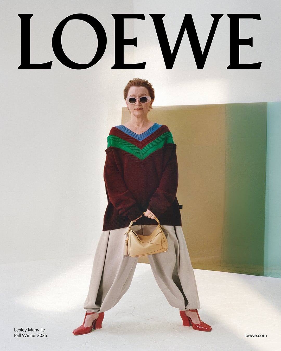 An elderly woman wearing sunglasses, a patterned knit sweater, pale trousers, and holding a handbag in a studio setting.